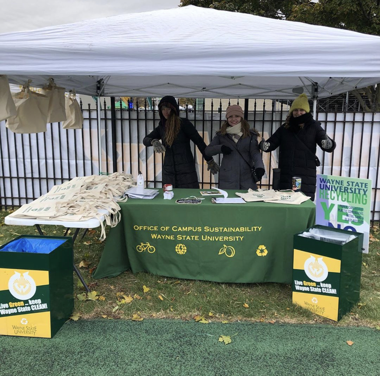 Three student interns in winter gear standing under a white canopy tent urging others to learn more about the Office of Campus Sustainability