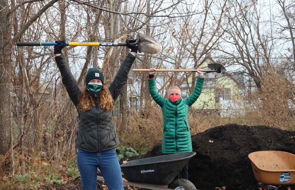 Students hold shovels high above their heads next to a large pile of compost.